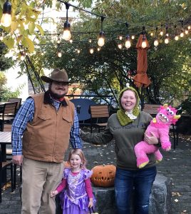 A man and woman stand in front of a pumpkin with two little girls in Halloween costumes.